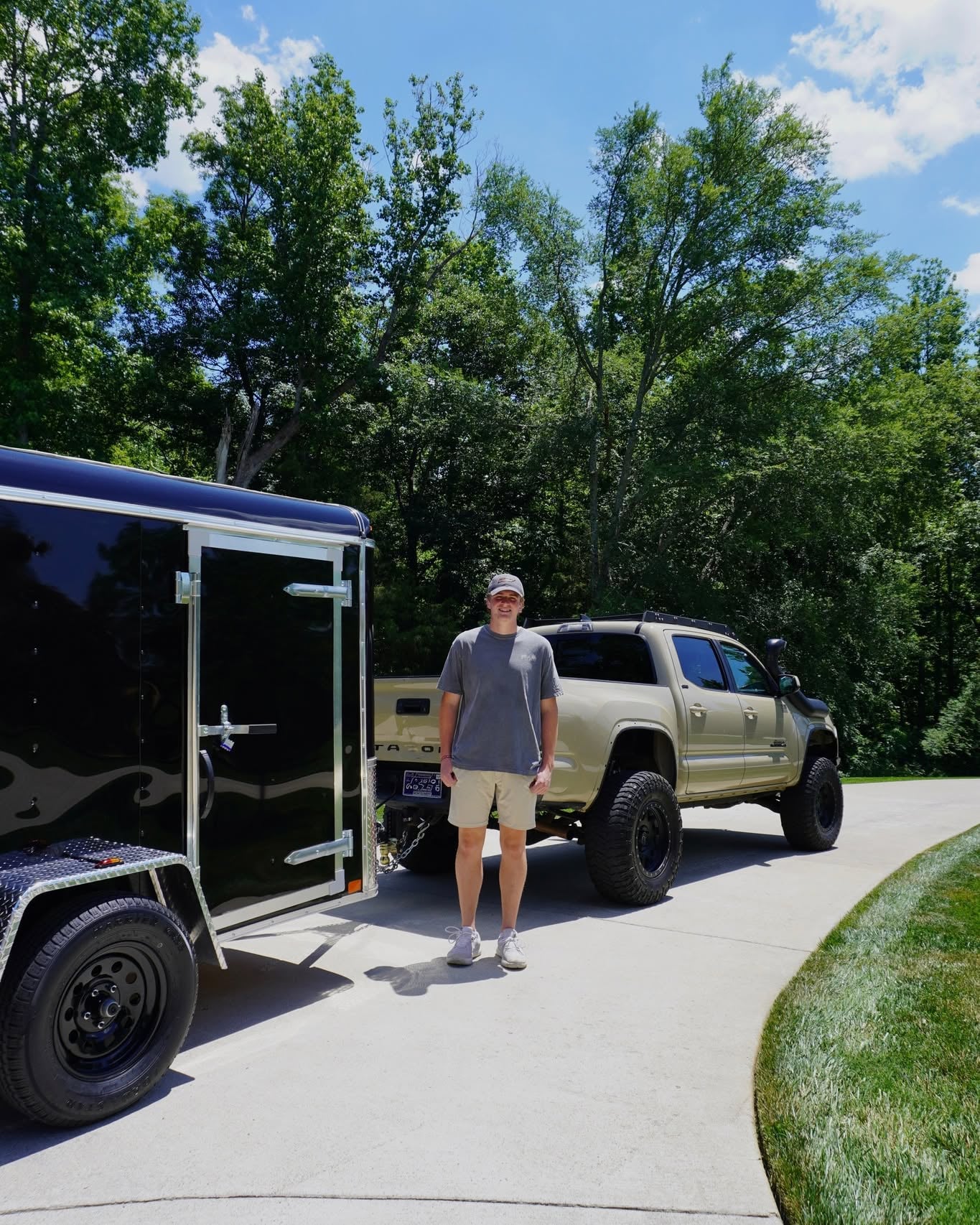 Sam Hoffman standing beside his tan Toyota Tacoma and black enclosed detailing trailer on a sunny driveway