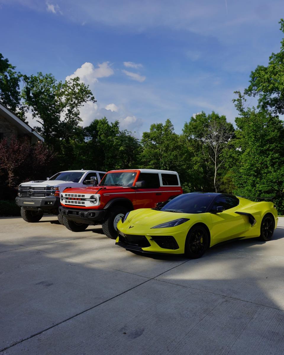 Three-vehicle exterior detail: white Chevy Silverado, red Ford Bronco, and yellow C8 Corvette lined up on a driveway after full exterior detail